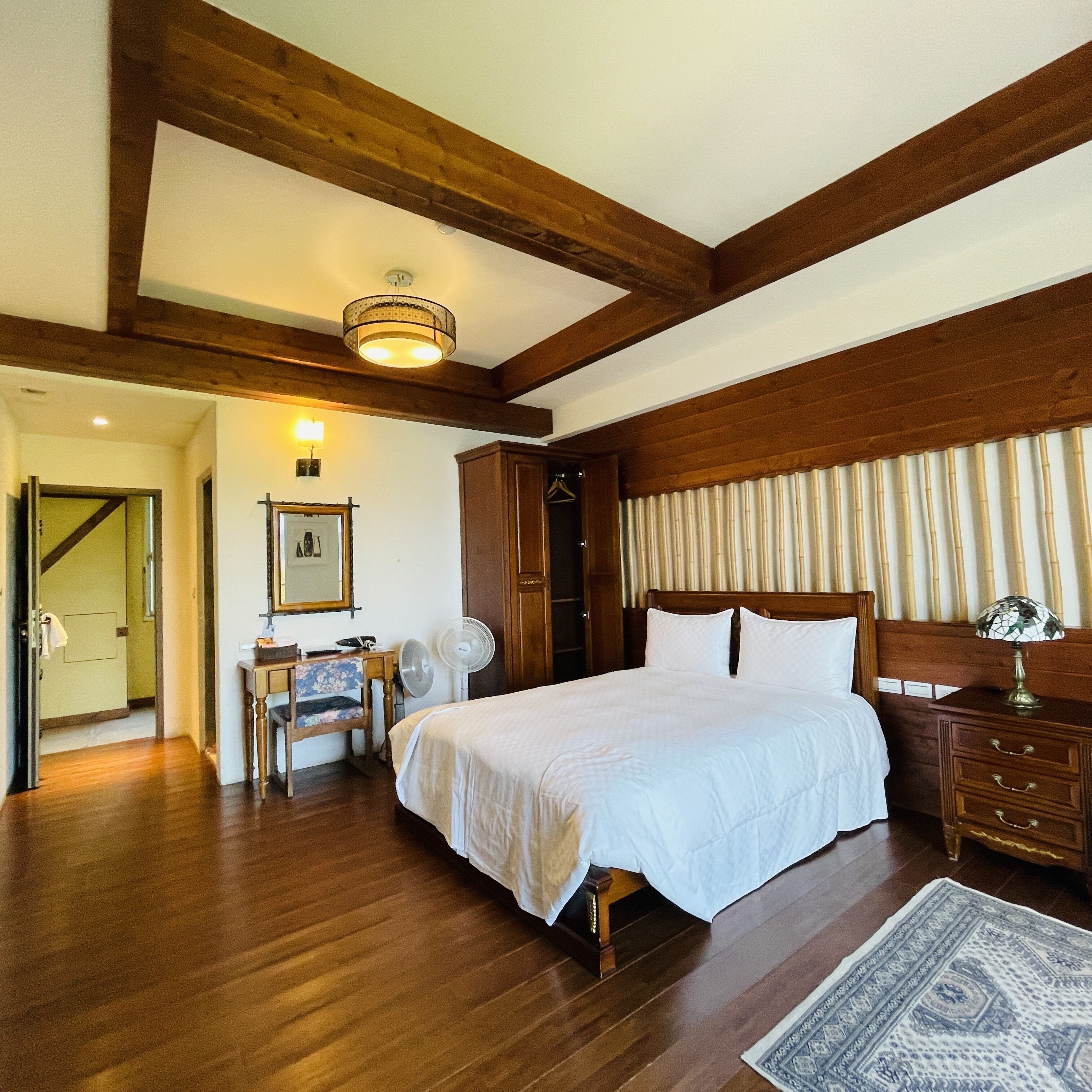 A bedroom with a wooden ceiling, a bed with white bedding, a bedside table with a lamp, and a chair.