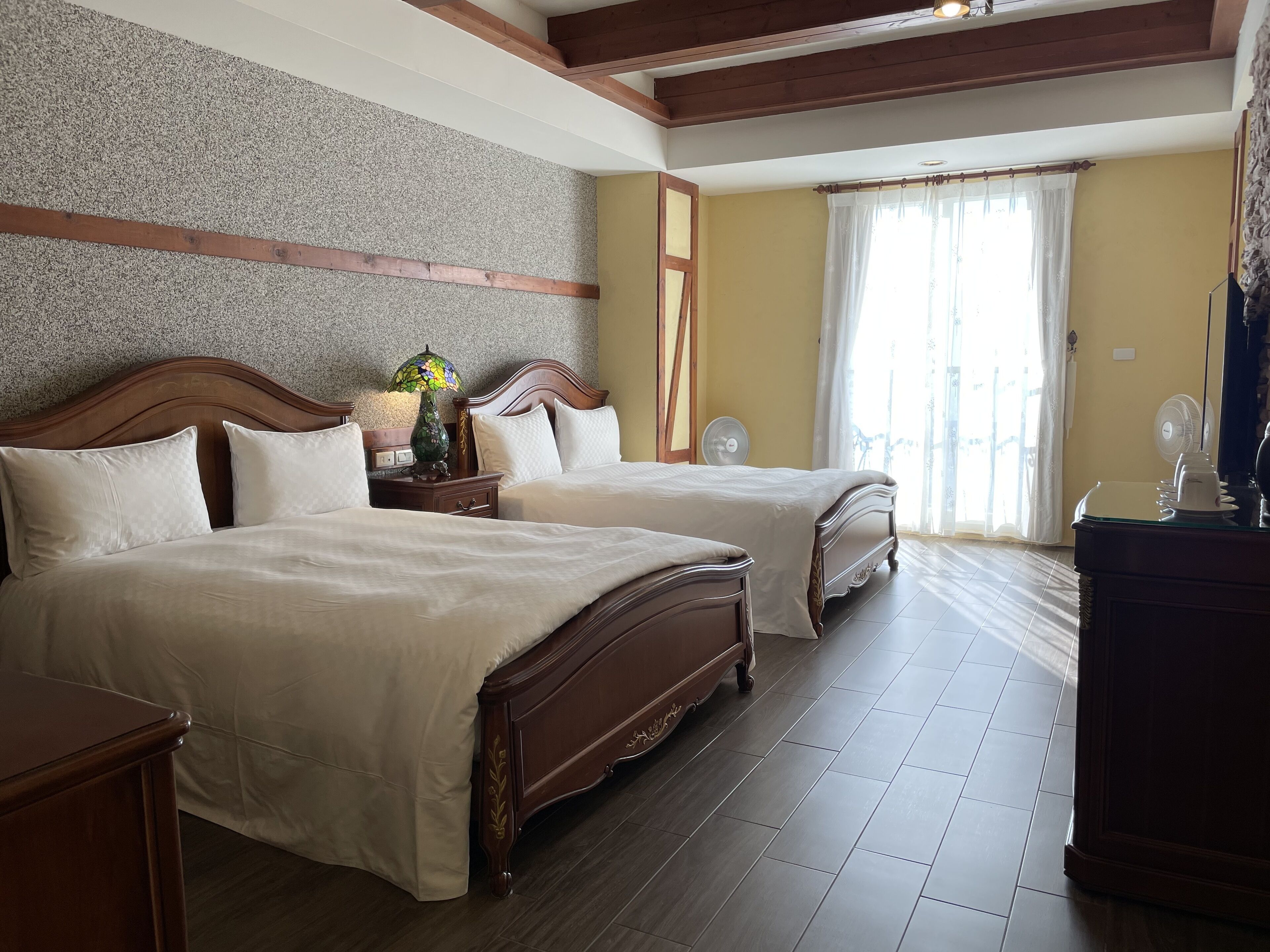 Habitación de hotel con dos camas, cabecera de madera, lámpara decorativa, ventilador y un ventanal con cortinas.