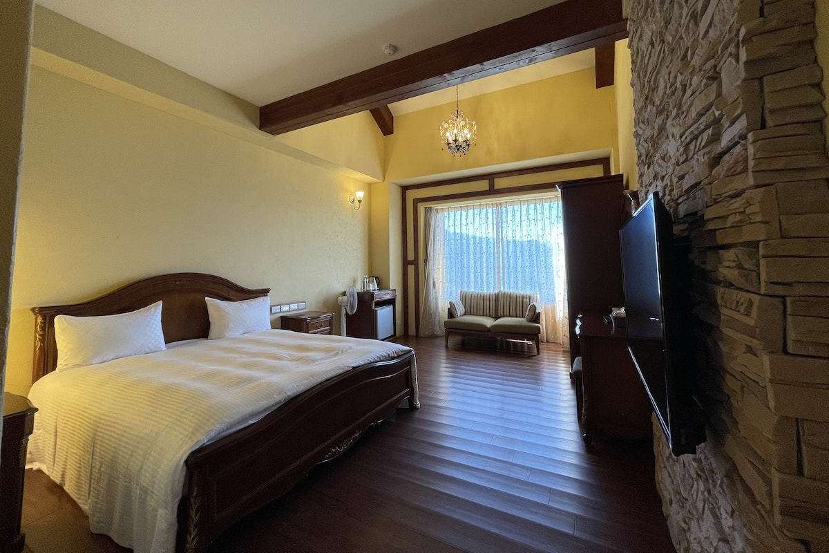 A bedroom with a large bed, a TV mounted on a stone wall, a wooden ceiling, and a stone fireplace.