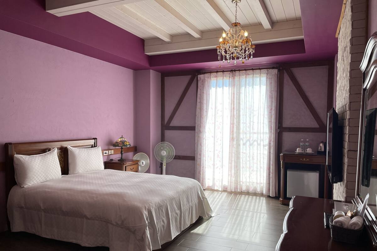 A bedroom with a large bed, a bedside table with a lamp, a fan, and a TV on a dresser.