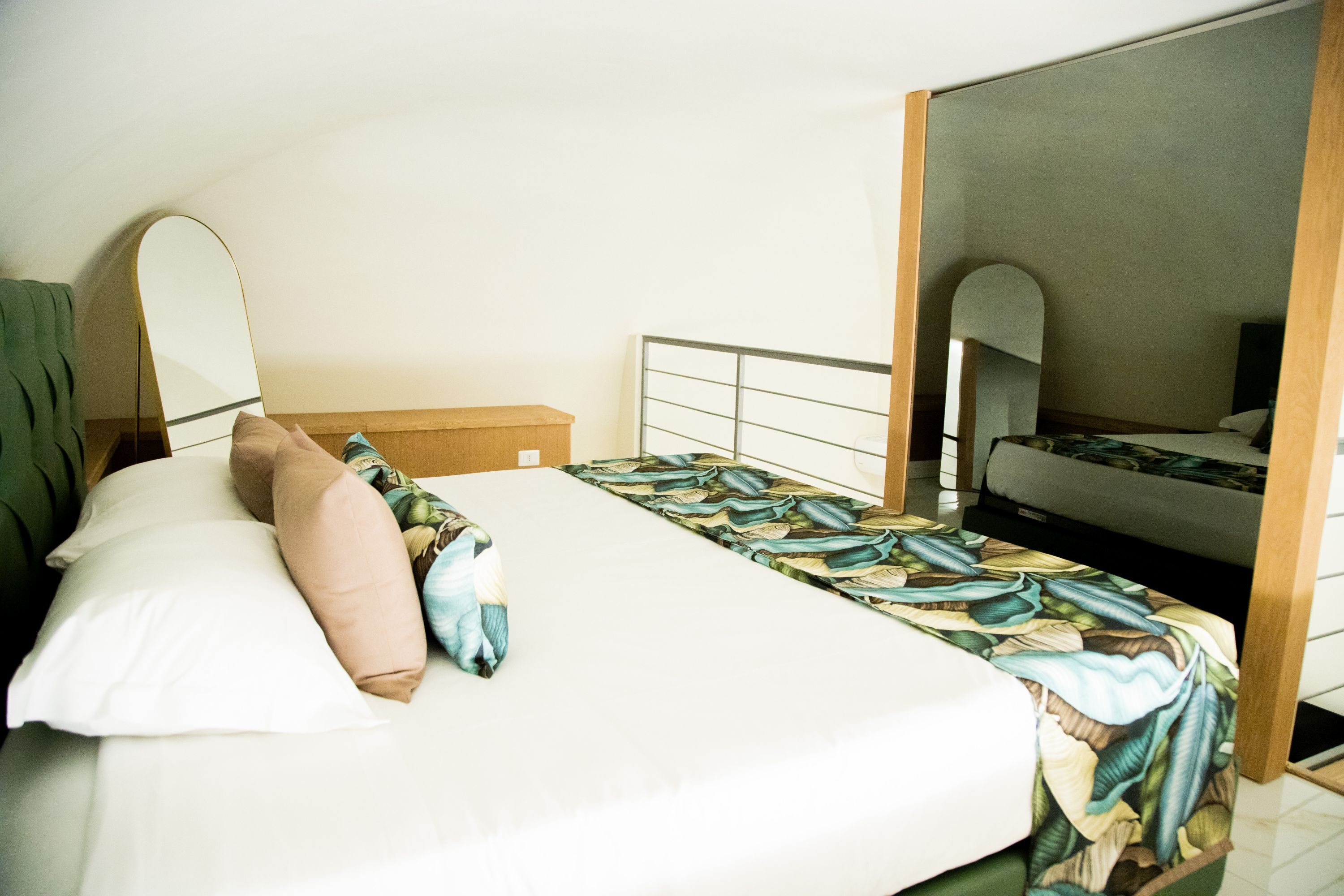A neatly made bed with a patterned bedspread and pillows, a wooden nightstand, a mirror, and a view of another room.