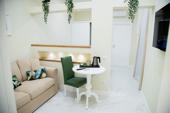 A small, well-lit room with a beige sofa, a green chair, a white table with a kettle, and a TV mounted on the wall.