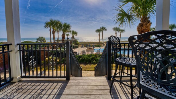 A covered outdoor area with a view of the ocean, palm trees, and a pool area.