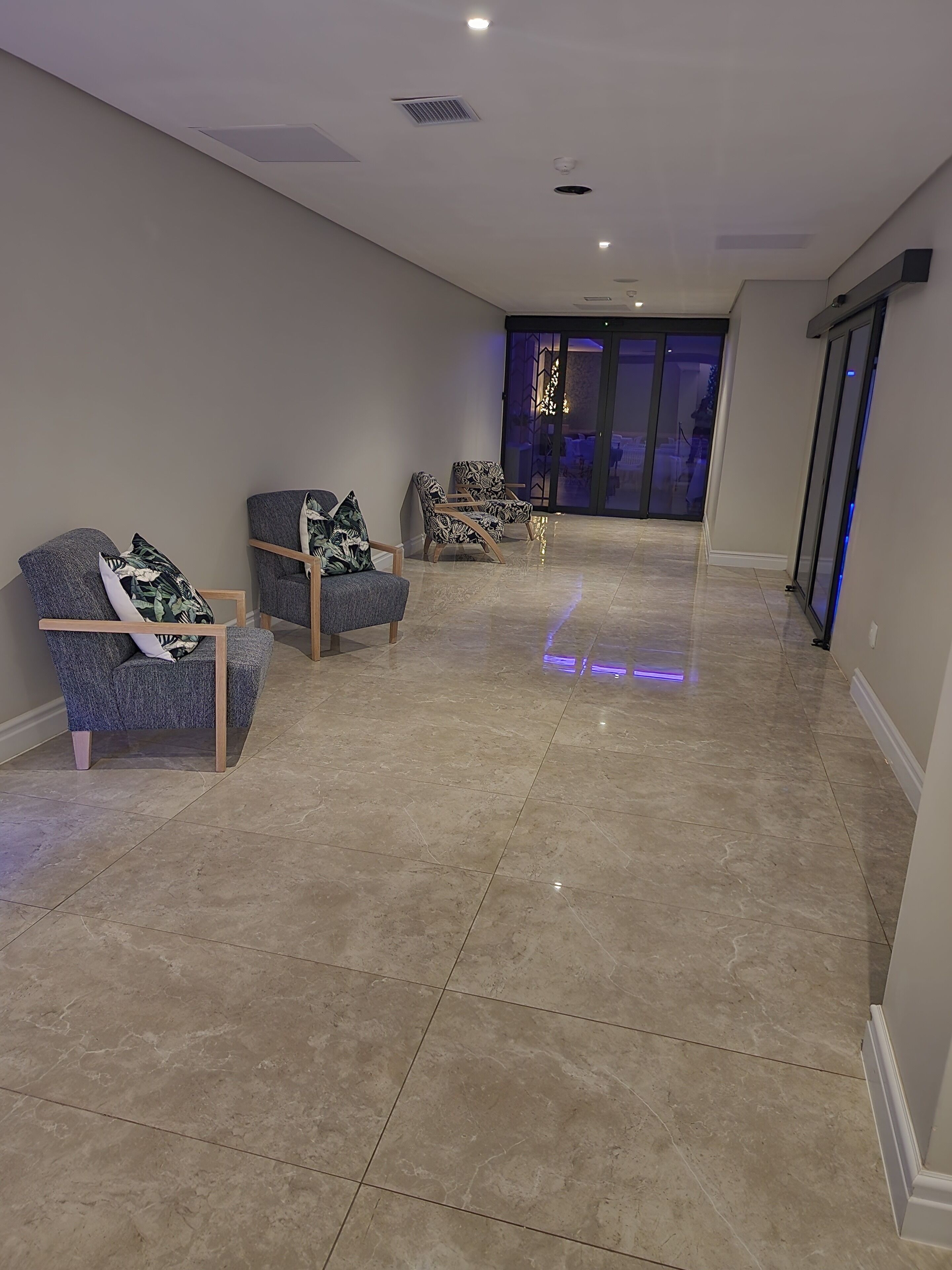 Un pasillo amplio con piso de baldosas, dos sillones con cojines estampados y un ventanal corredera que da a un espacio exterior.