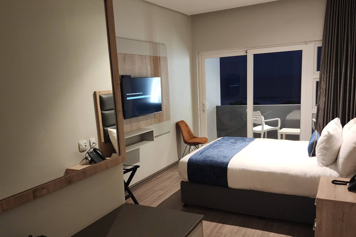 A modern hotel room with a bed, a desk, a chair, a TV, and a balcony with a view.