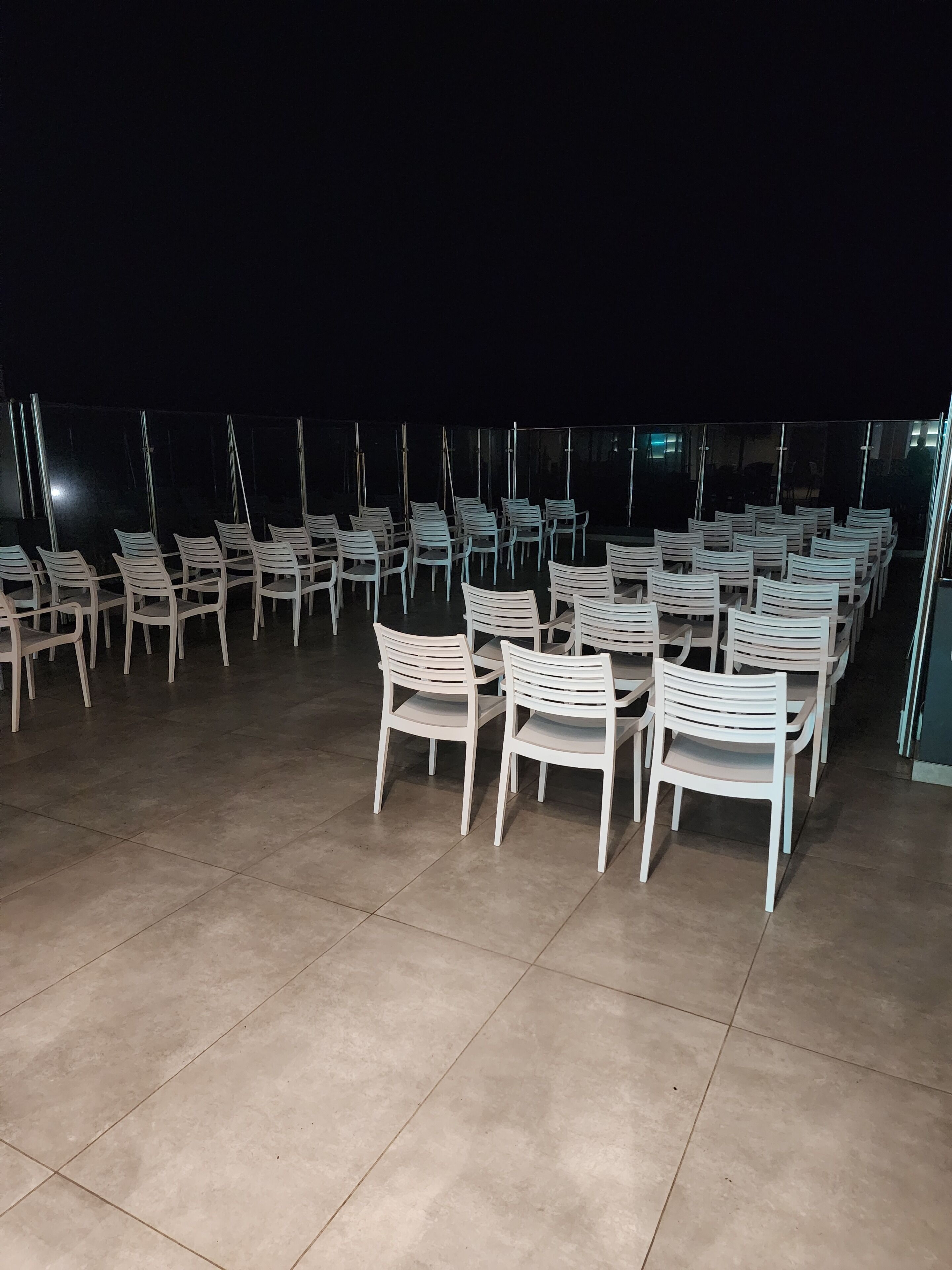 An outdoor seating area with rows of white plastic chairs and tables, arranged on a tiled floor.