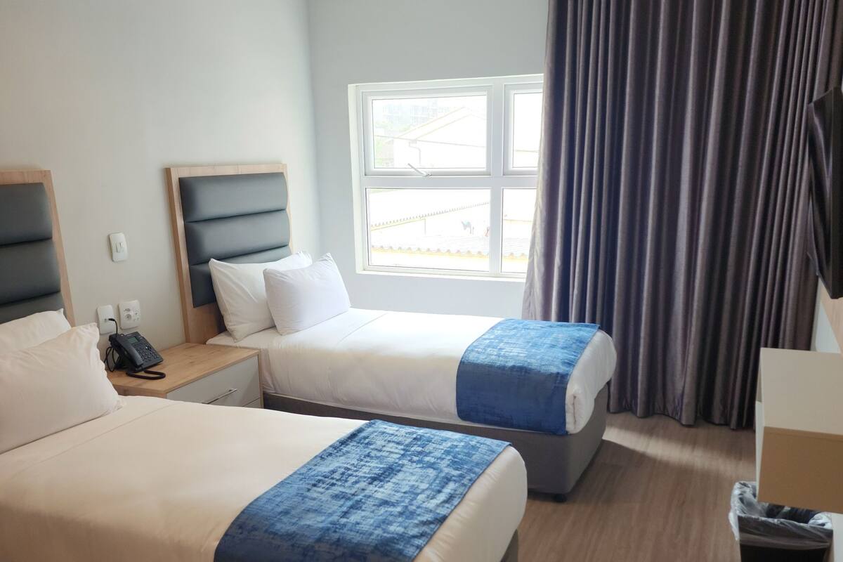A hotel room with two beds, a window with curtains, a bedside table with a phone, and a trash bin.