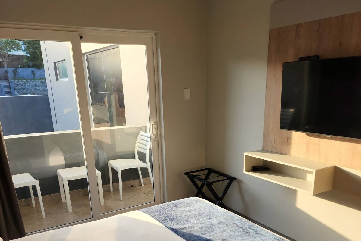 A hotel room with a bed, a TV mounted on the wall, a small table, and a balcony with chairs.
