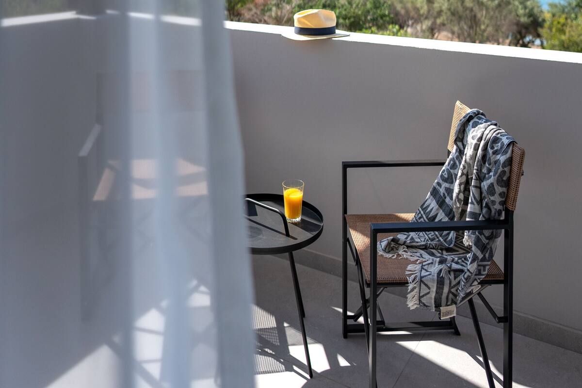 A balcony with a view of the sea, a table with a glass of orange juice, and a chair with a patterned throw.