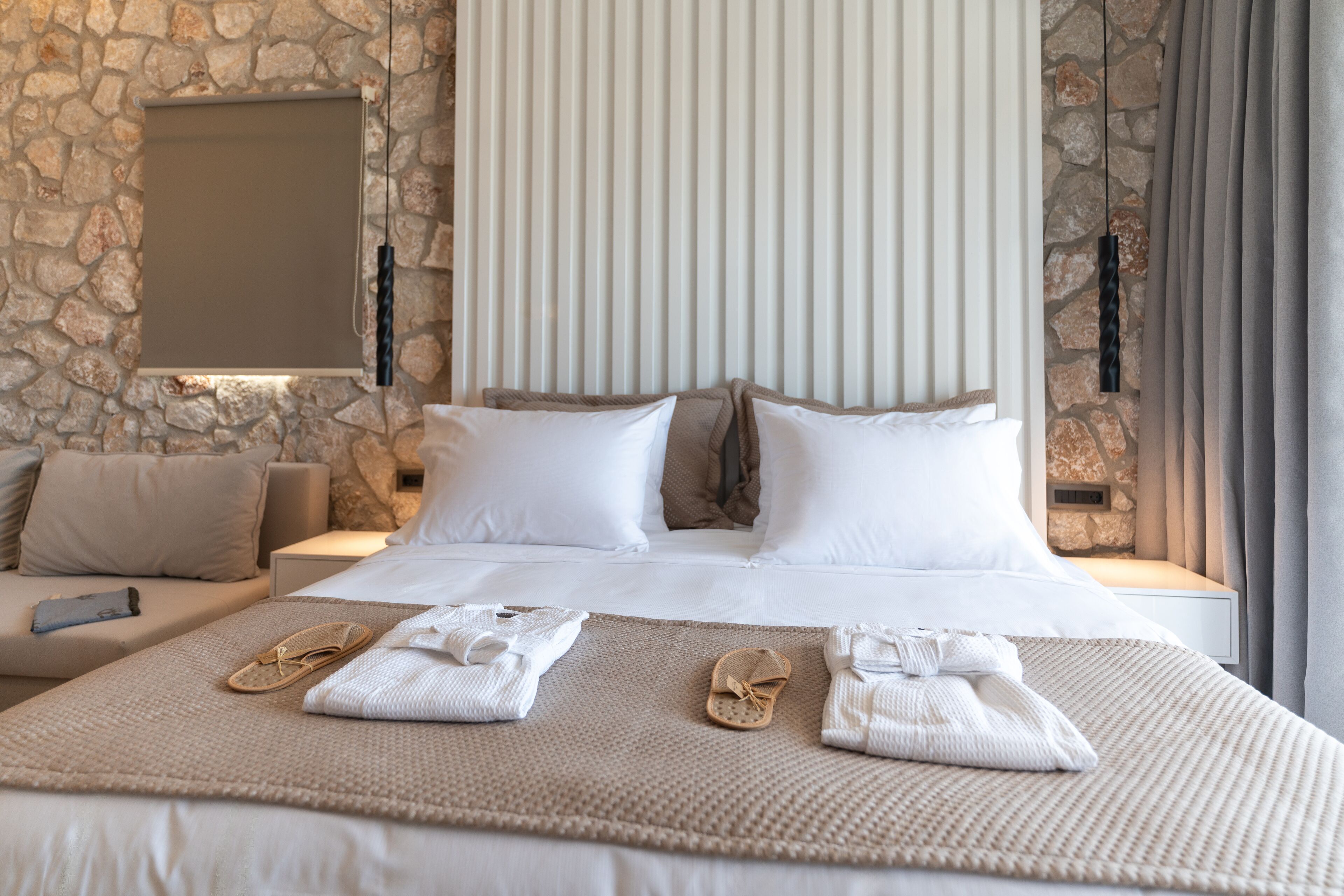 A well-appointed hotel room with a stone wall, a large bed with white linens, a sofa, and a flat-screen TV mounted on the wall.