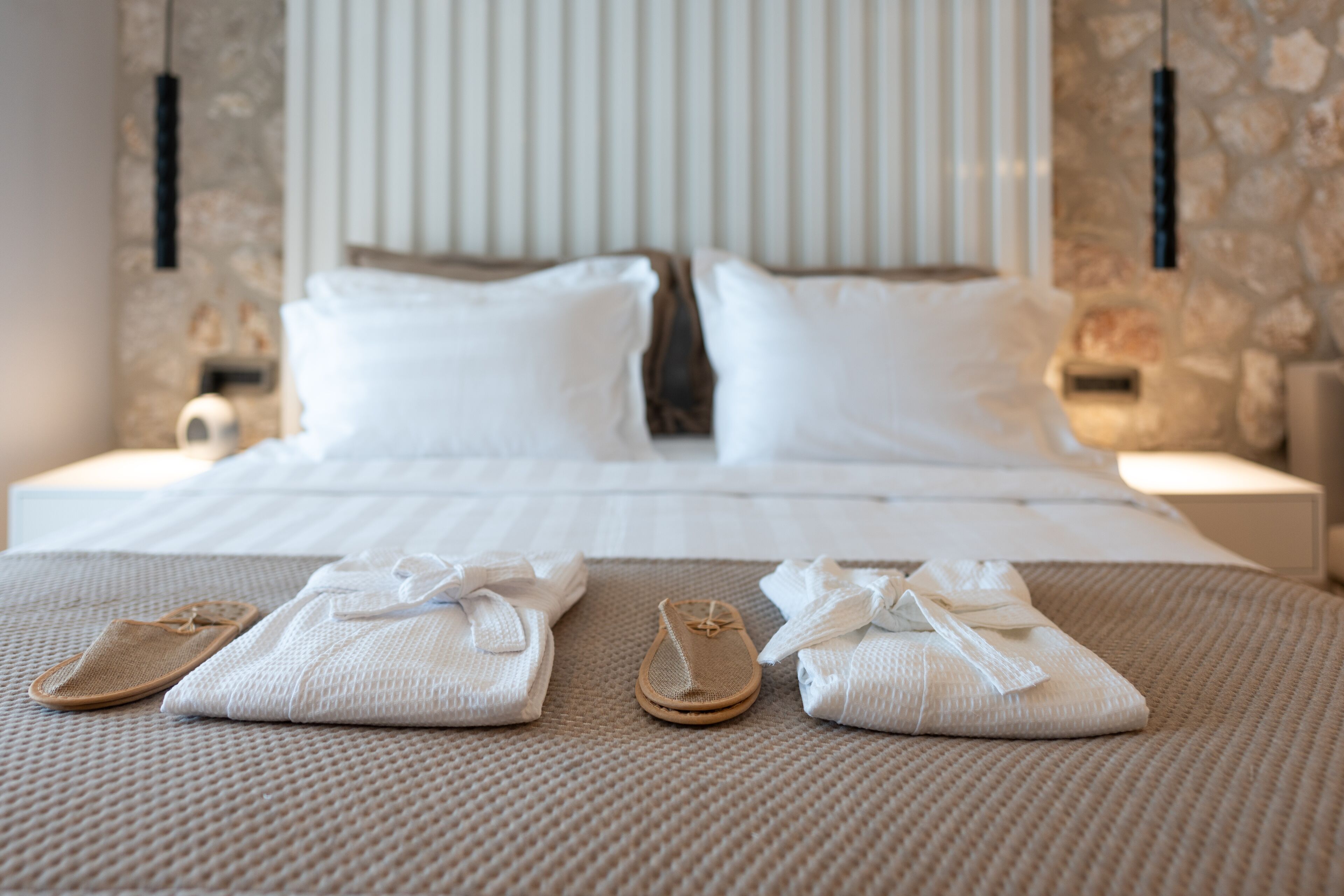 Una cama bien hecha con sábanas blancas, un par de pantuflas y toallas sobre un edredón texturizado.