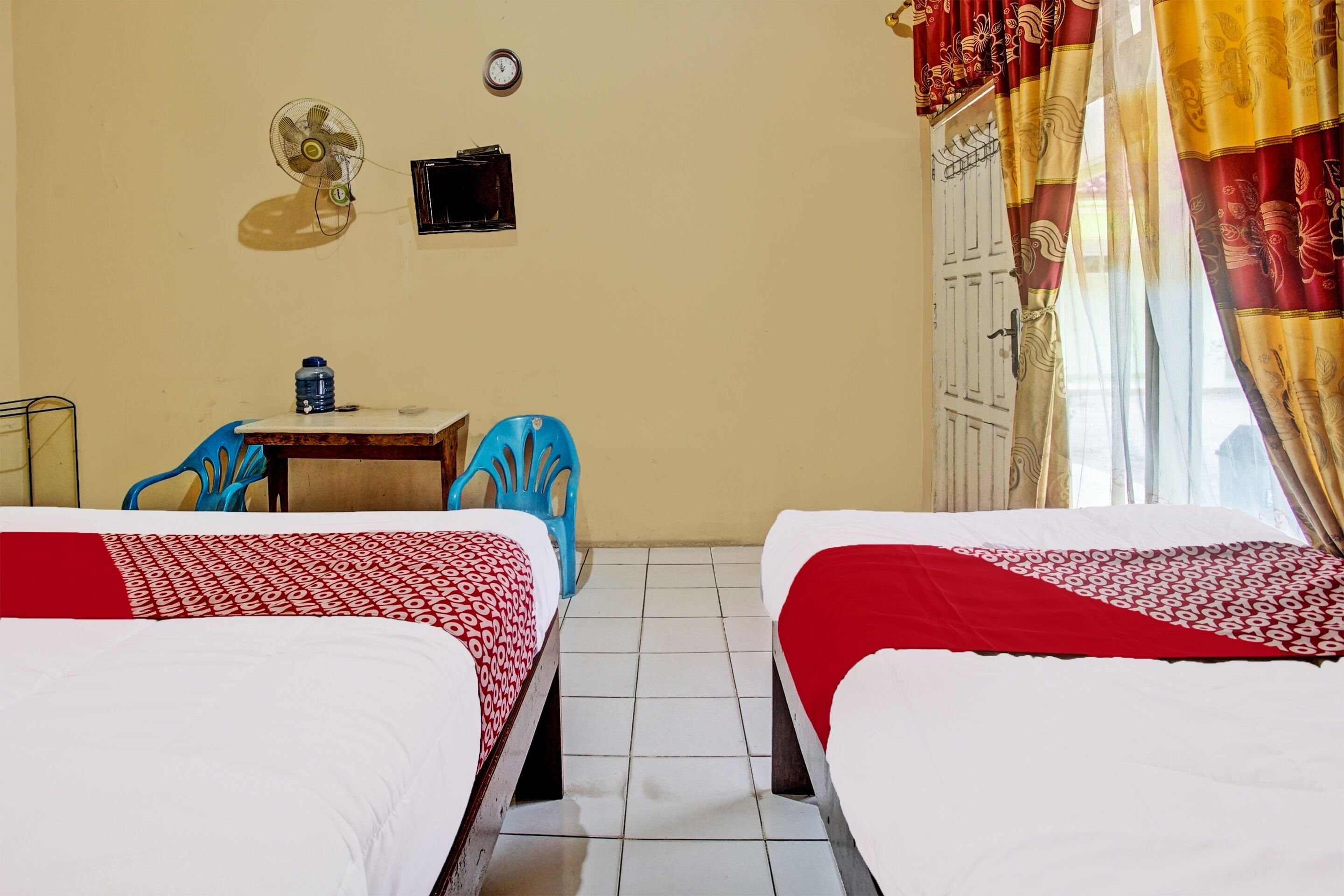 A room with two beds, a desk with a chair, a fan, a clock, and a wardrobe.