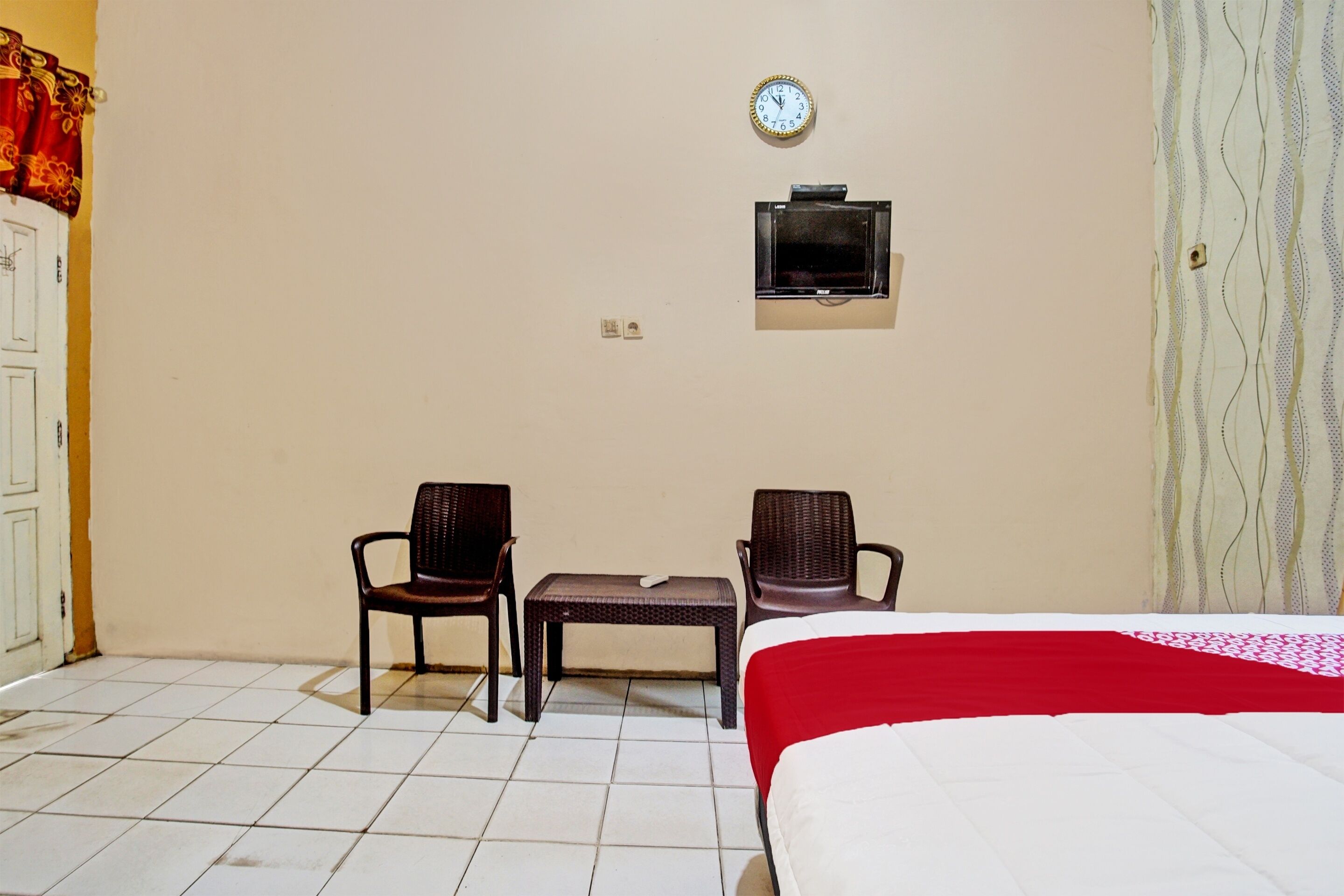 A hotel room with a bed, two chairs, a small table, a clock, and a television on the wall.