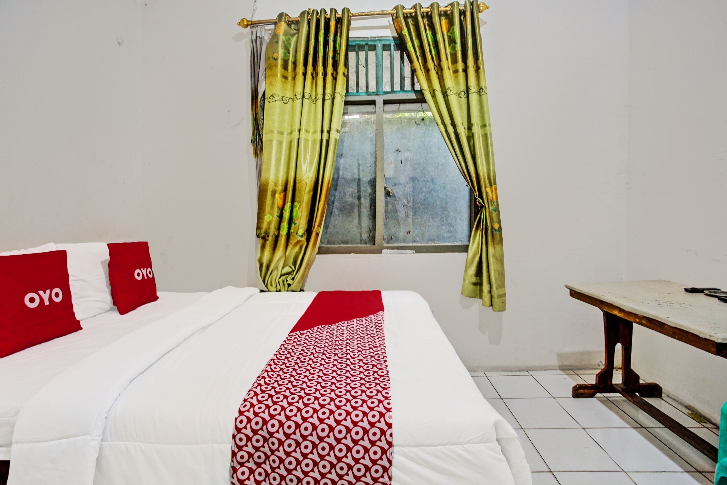 A room with a bed, two red pillows, a patterned blanket, a window with yellow curtains, and a wooden table.