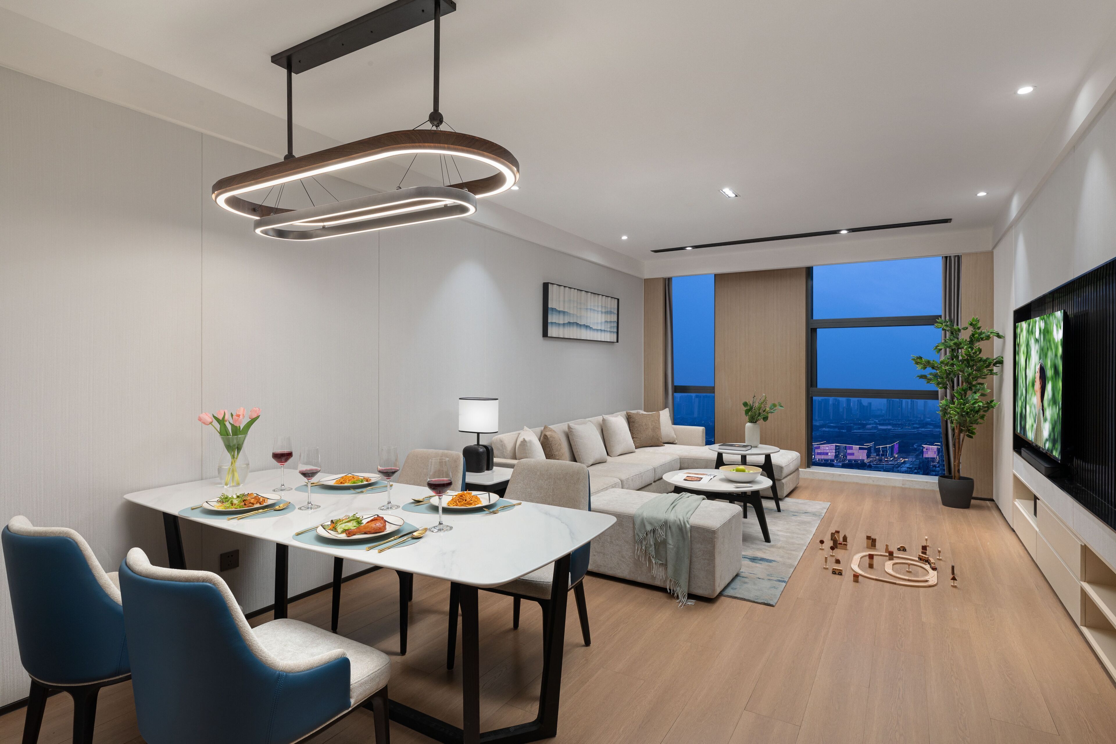 A modern living room with a dining area, a sofa set, a flat-screen TV, and a large window with a city view.