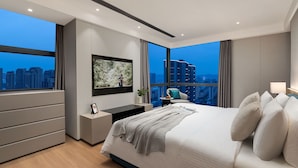 A modern hotel bedroom with a large bed, a flat-screen TV, a nightstand, and a view of the cityscape. - Somerset Taihu New City CBD Wuxi (Wuxi)