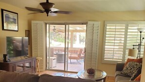 Un salon comprenant un canapé, un téléviseur et un ventilateur de plafond. Une porte-fenêtre coulissante donne accès à un patio extérieur avec une table et des chaises.