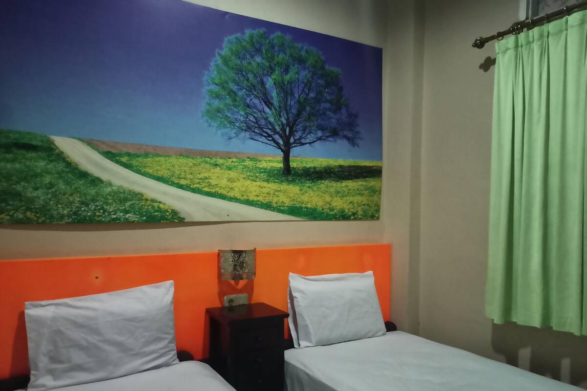 Une chambre avec deux lits, une tête de lit orange, une table de chevet, un rideau vert et un tableau représentant un arbre accroché au mur.