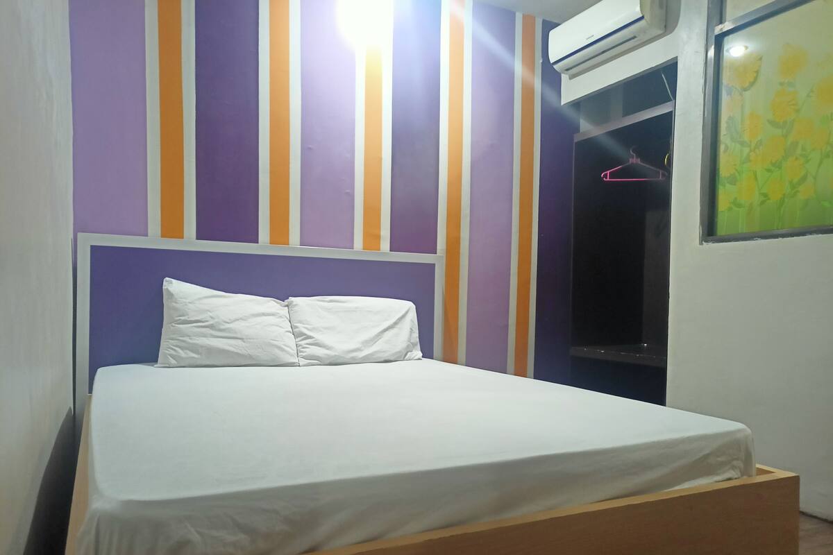 Un lit avec un cadre en bois, une literie blanche et une tête de lit adossée à un mur orné de rayures verticales violettes, oranges et blanches.