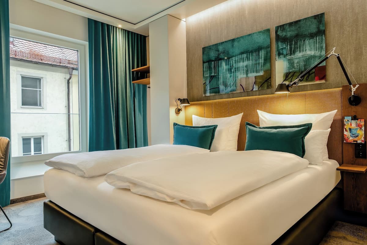 Una habitación de hotel con una cama grande, dos almohadas, una mesita de noche, una lámpara y una ventana con cortinas.