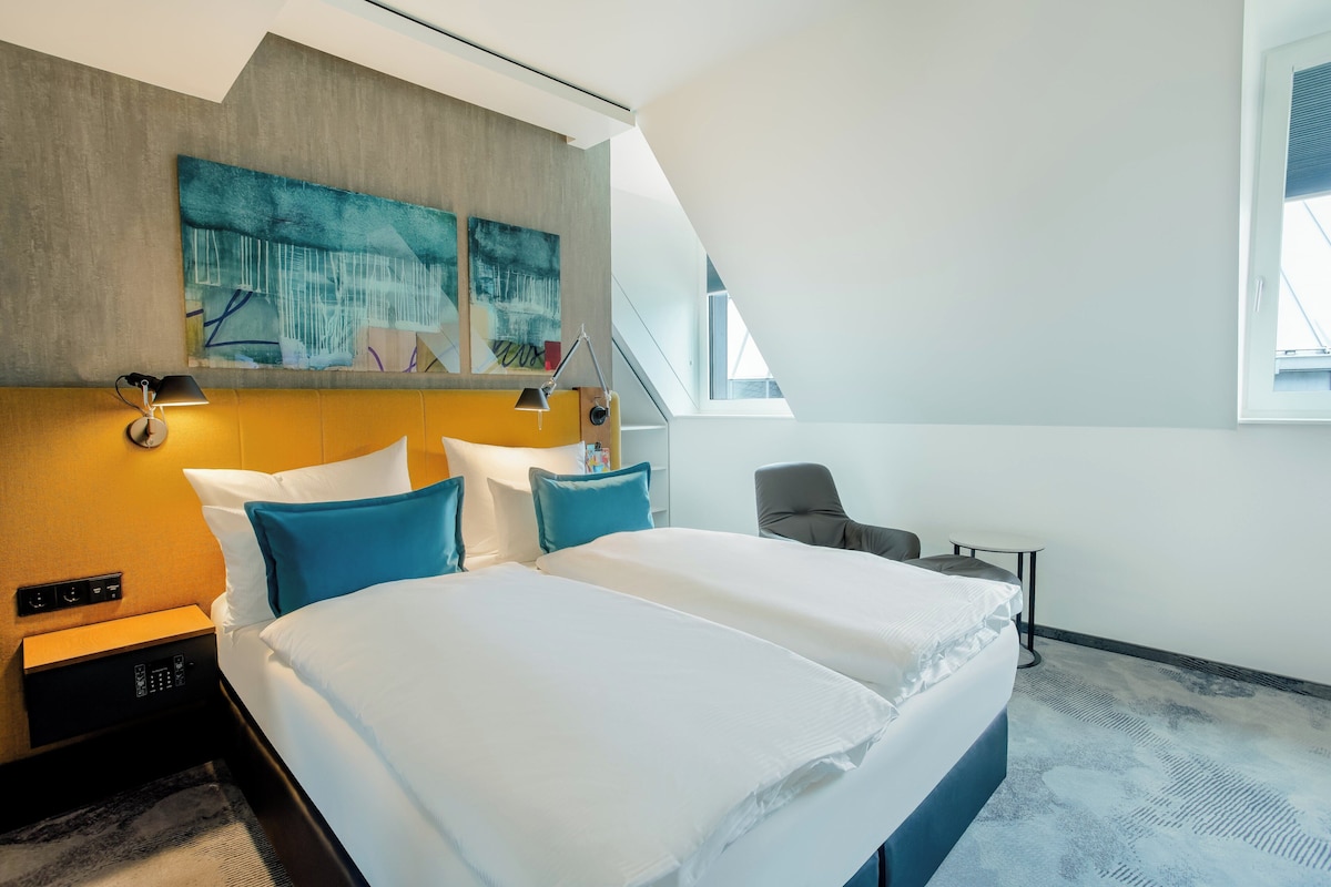 Ett modernt hotellrum med en stor säng, ett skrivbord och en stol. Det finns en tavla på väggen och ett fönster med utsikt över omgivningen.