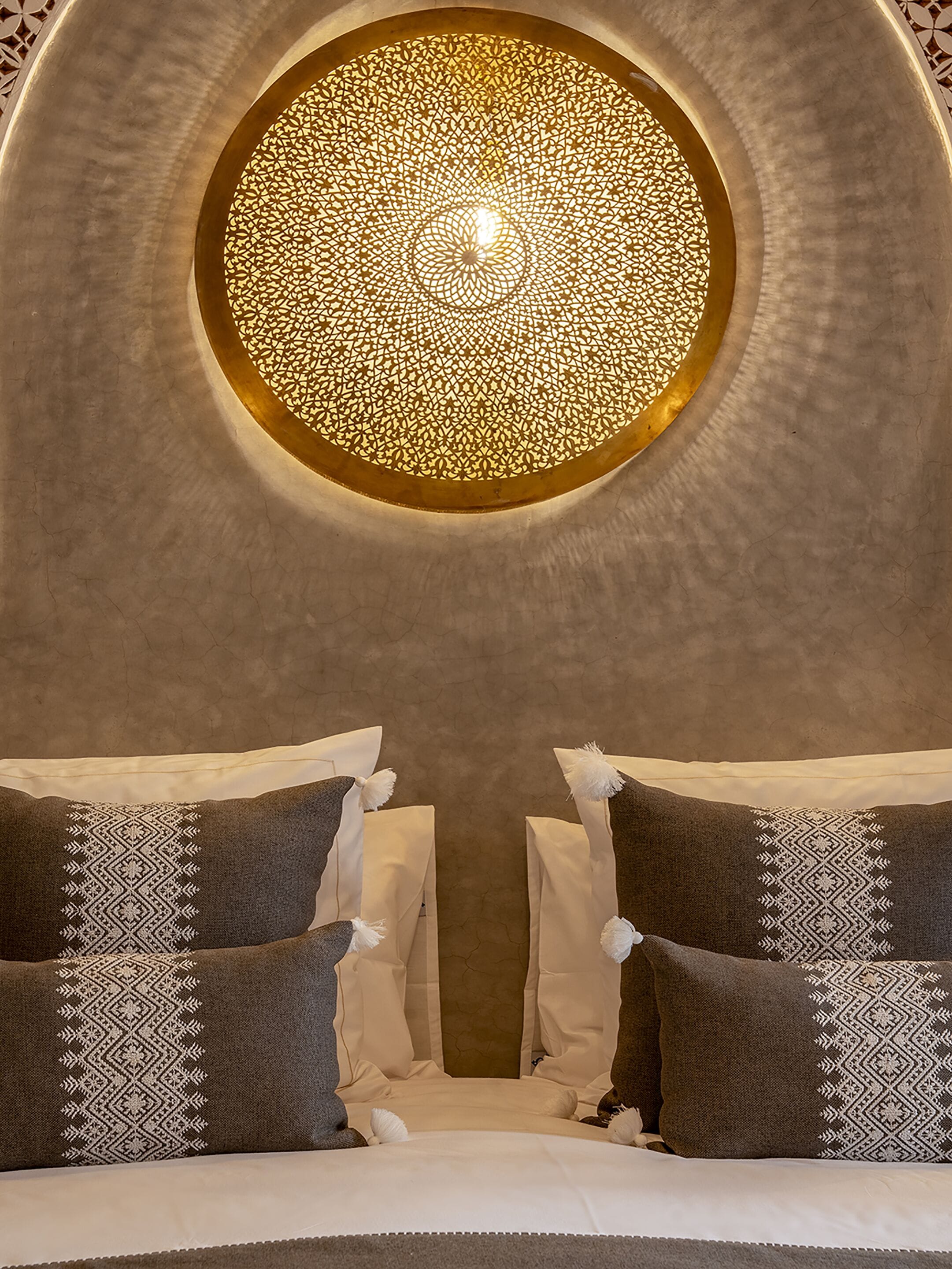 Una habitación con tragaluz circular, cama con almohadas estampadas y pared con textura.