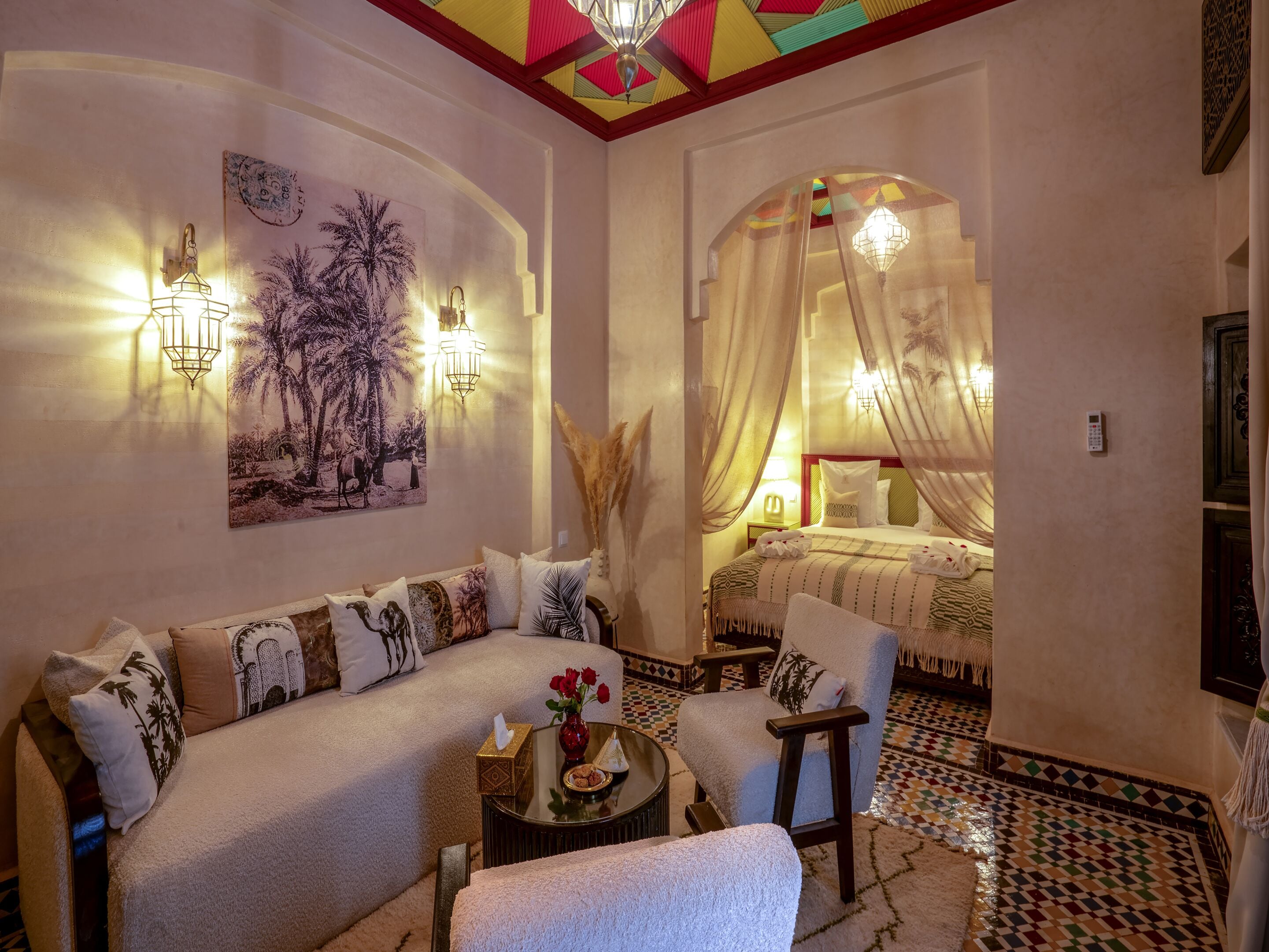 Una habitación acogedora con sofá, sillón y una cama con dosel. Hay luces fijadas en la pared y un cuadro de palmeras.