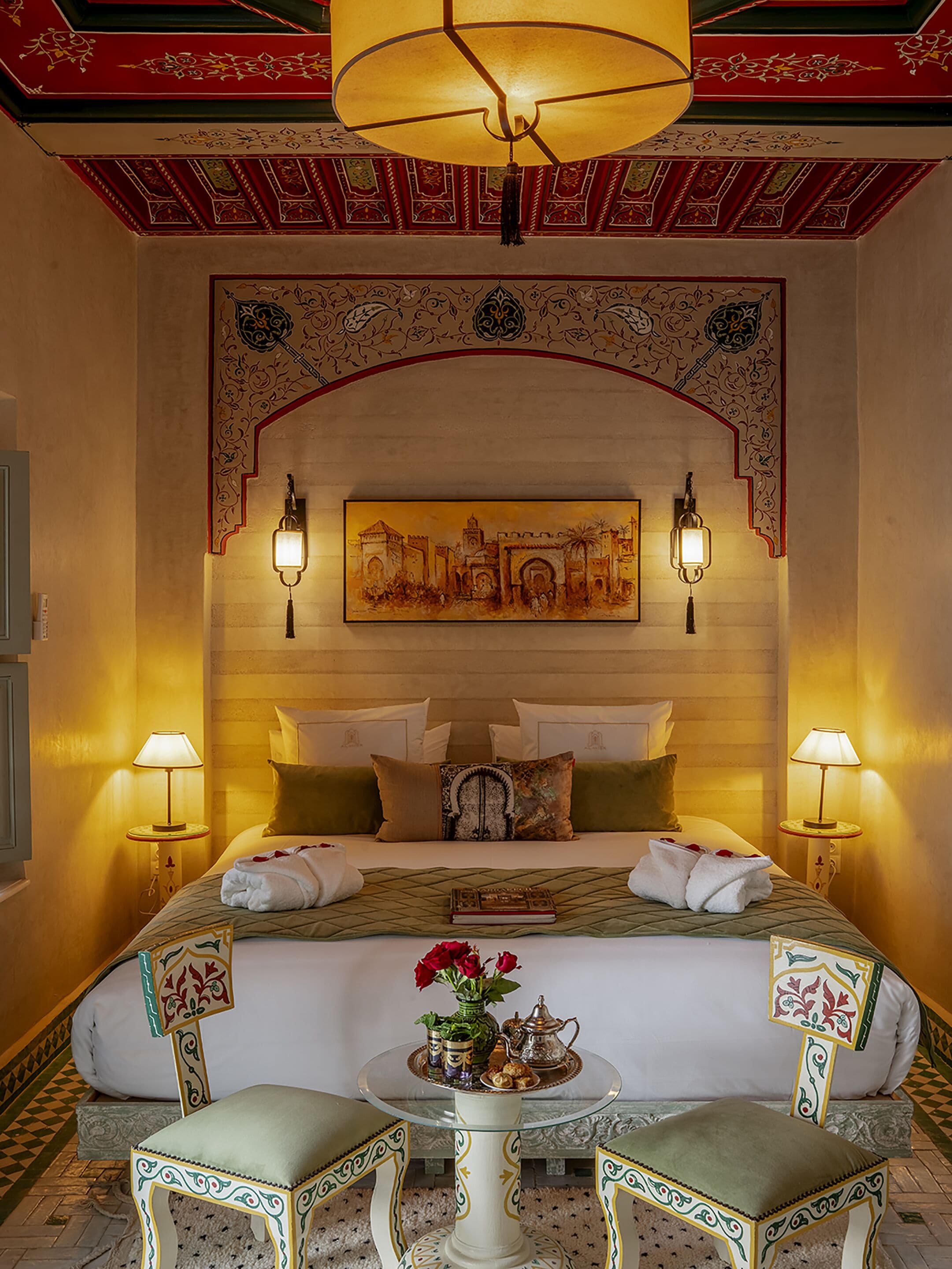 A hotel room with a large bed, two bedside lamps, a decorative wall piece, a small table with a vase and a book, and two chairs.