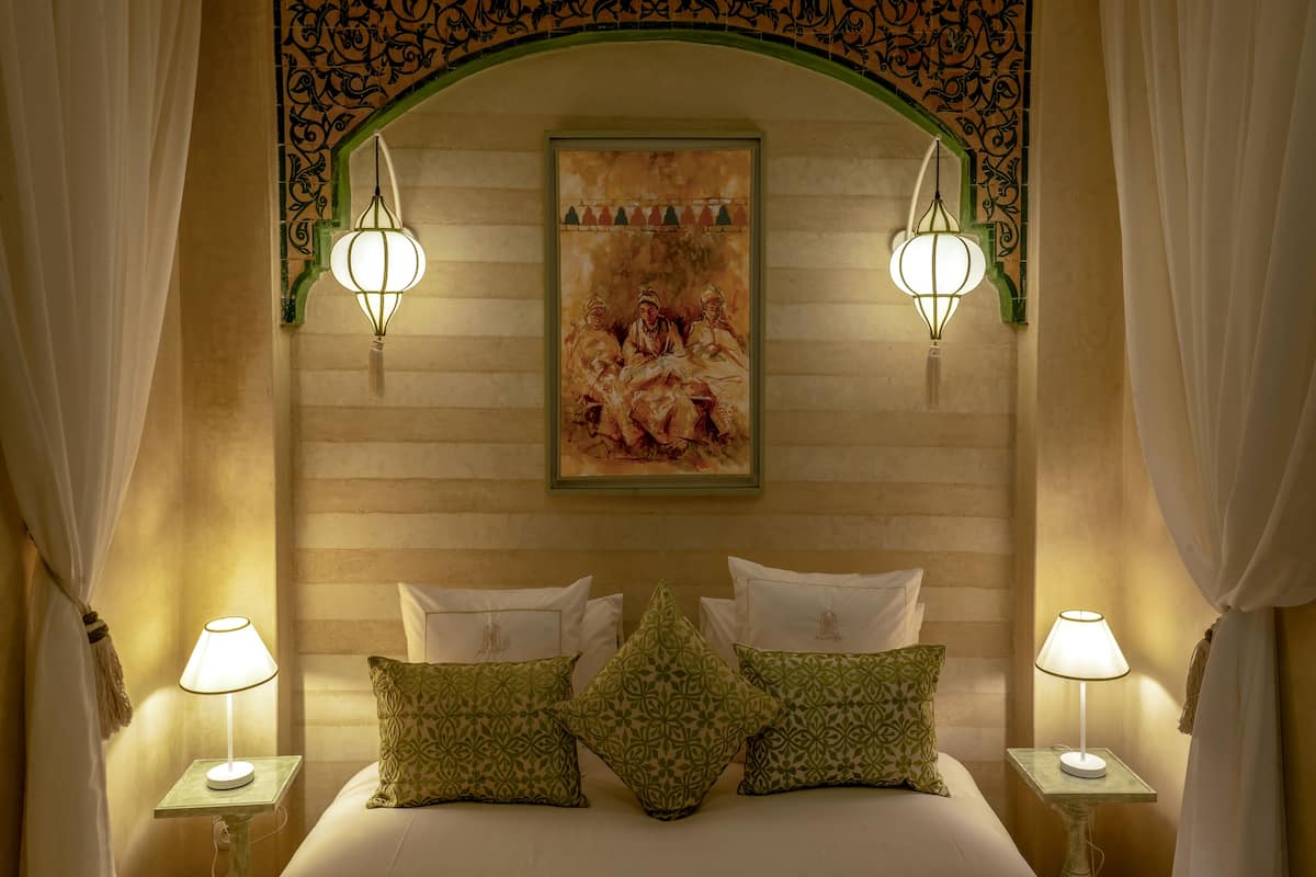 A bedroom with a bed, two bedside lamps, a painting on the wall, and draped curtains.