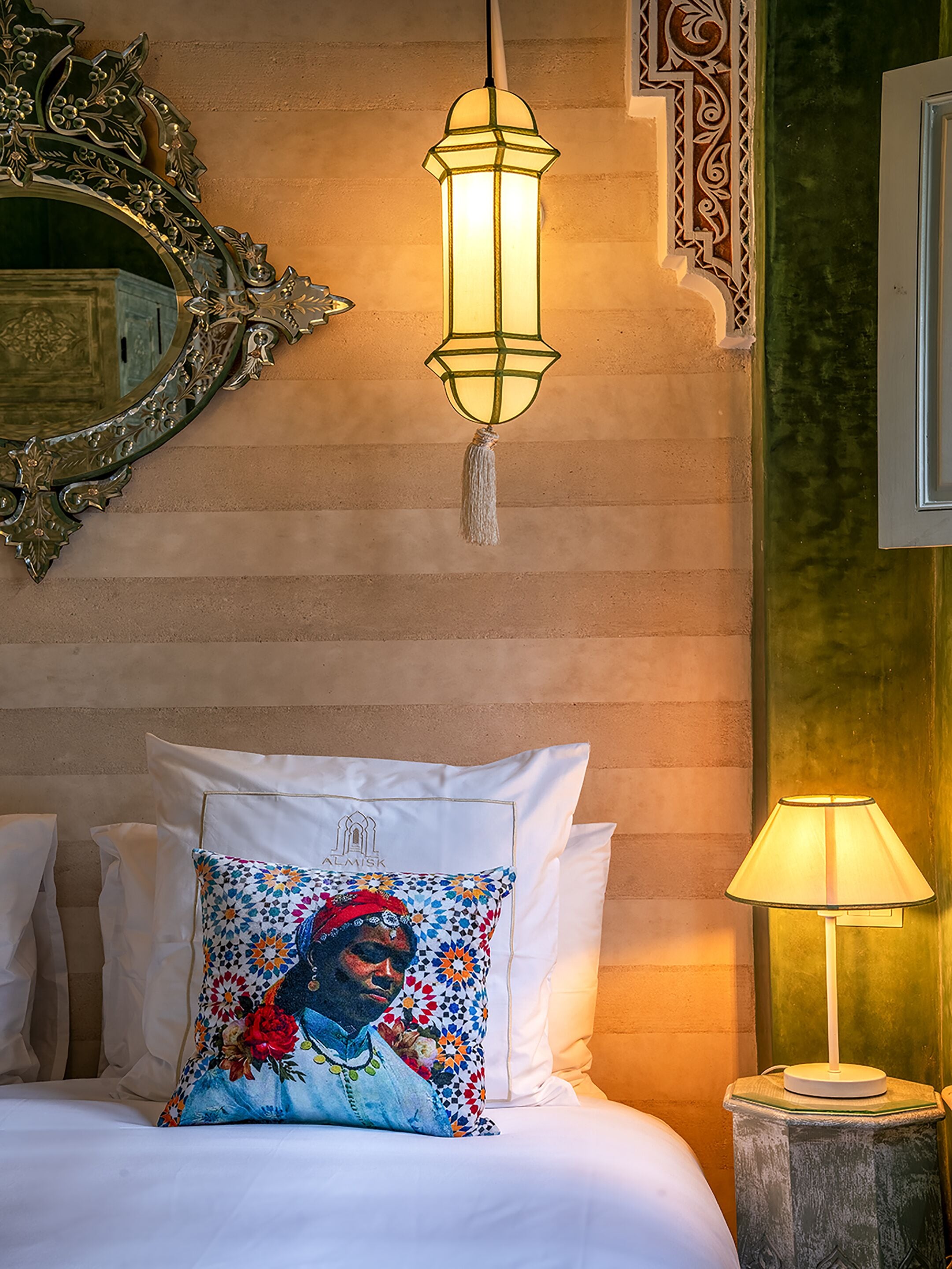 A bed with white linens, a colorful pillow, a decorative mirror, a wall-mounted light fixture, and a bedside table with a lamp.