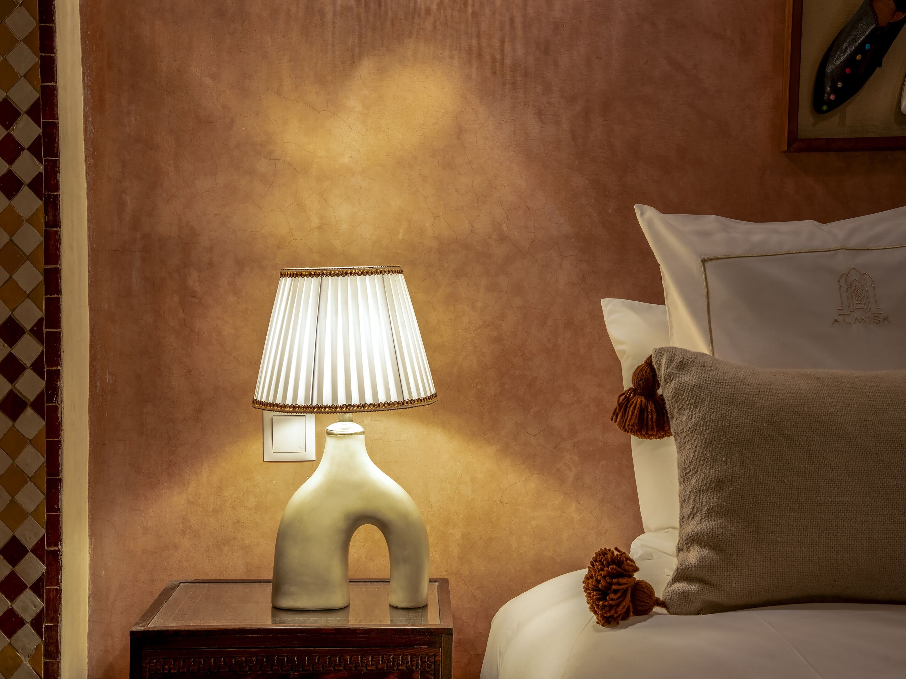 A neatly made bed with a white pillow and a decorative pillow, a bedside lamp, and a small wooden nightstand.