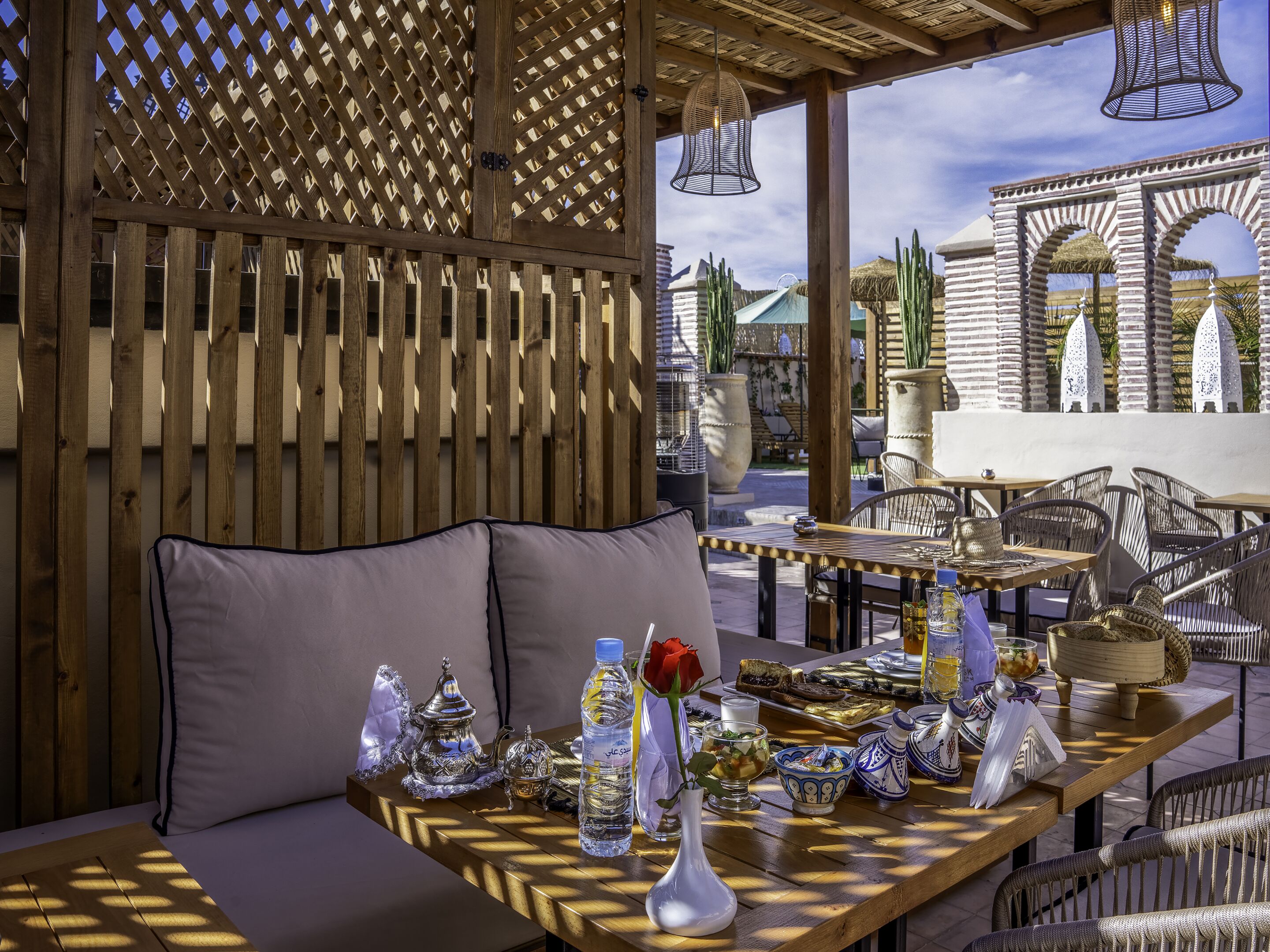 An outdoor seating area with wooden lattice walls, cushioned seating, and tables set with various items including a water bottle, a rose, and decorative bowls.