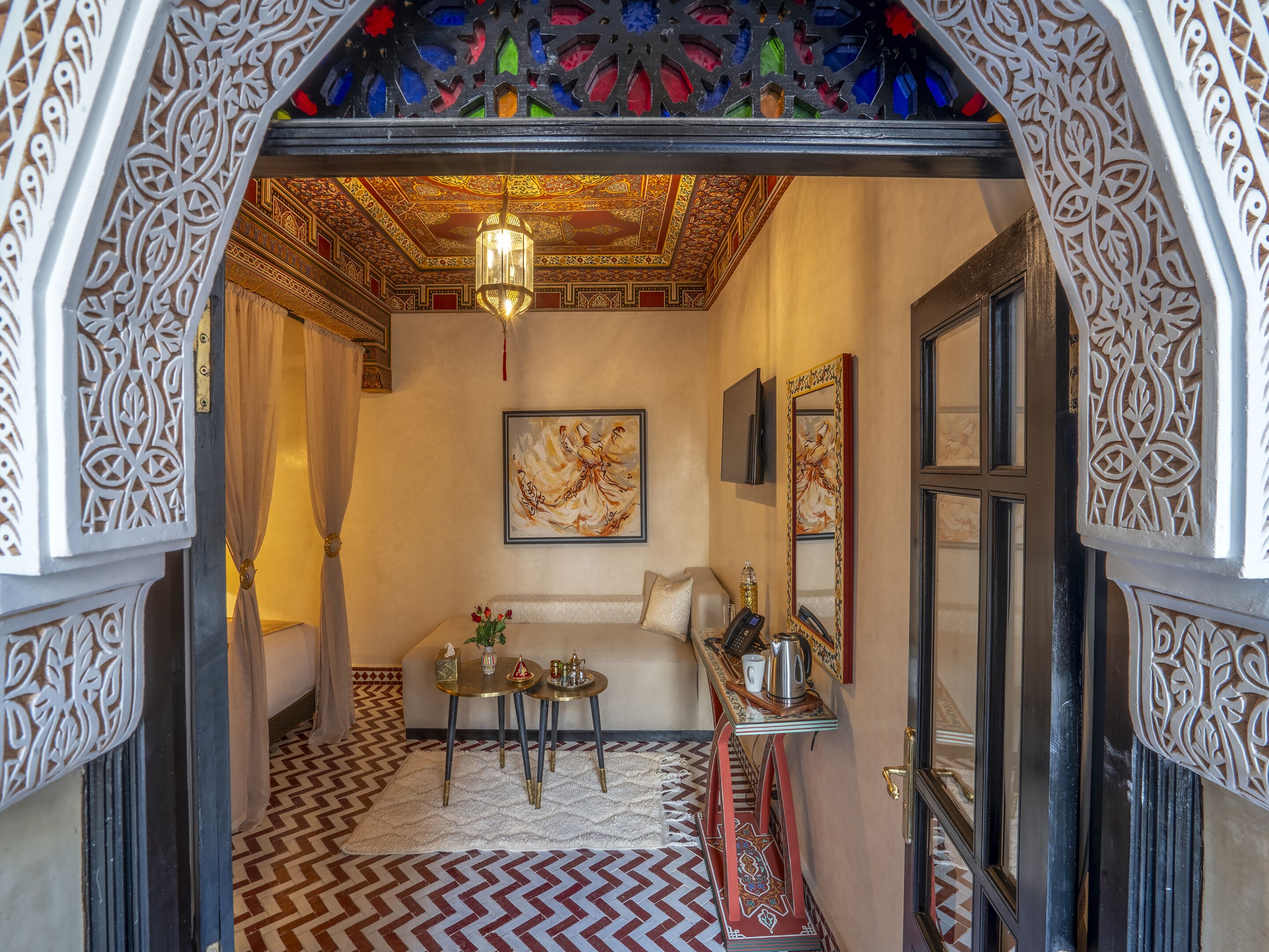 A room with a patterned floor, a bed, a small table with a vase, a wall-mounted TV, and a decorative ceiling.