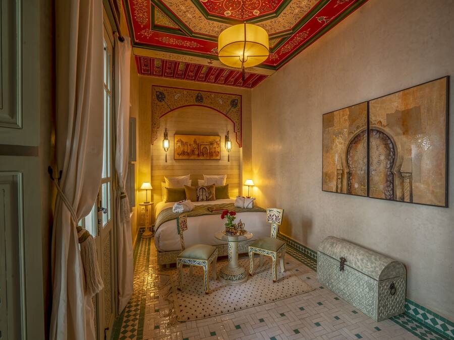 Una habitación tradicional marroquí con una cama, dos mesitas de noche, una pequeña mesa y un cofre decorativo.