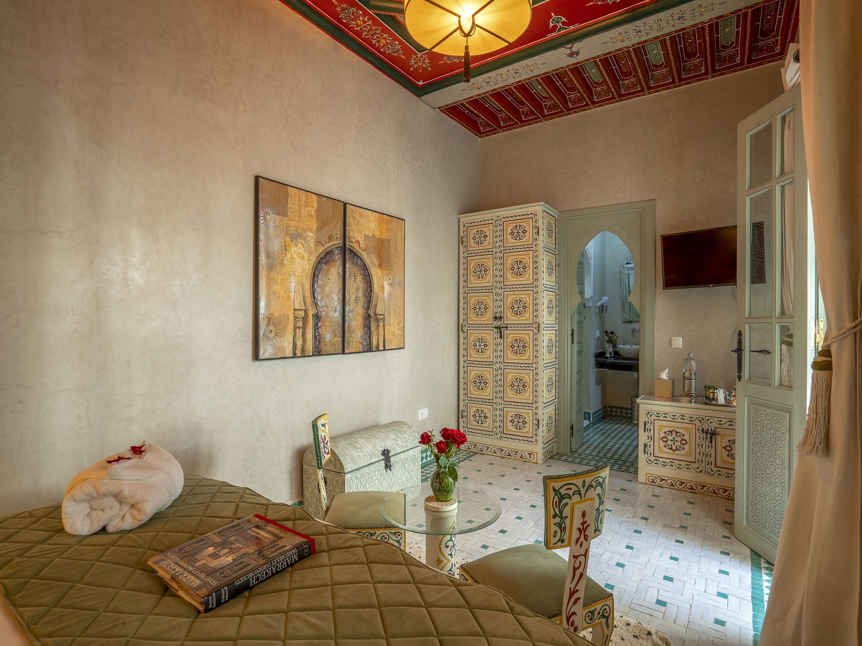 Una habitación con una cama, un adorno en la pared, una mesa de vidrio con un florero y vista a otra habitación con un espejo y un televisor.