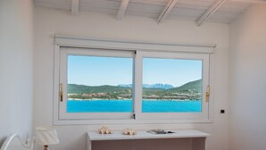 Quarto com cama, mesa de cabeceira, luminária, janela com vista para o mar e montanhas, e uma pequena mesa com objetos decorativos.