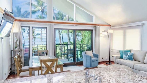 A spacious living area with a large sliding glass door leading to a balcony, a dining table with chairs, a sofa, and a floor lamp. - Beachfront luxury | Walk to dining & shops (Kihei)