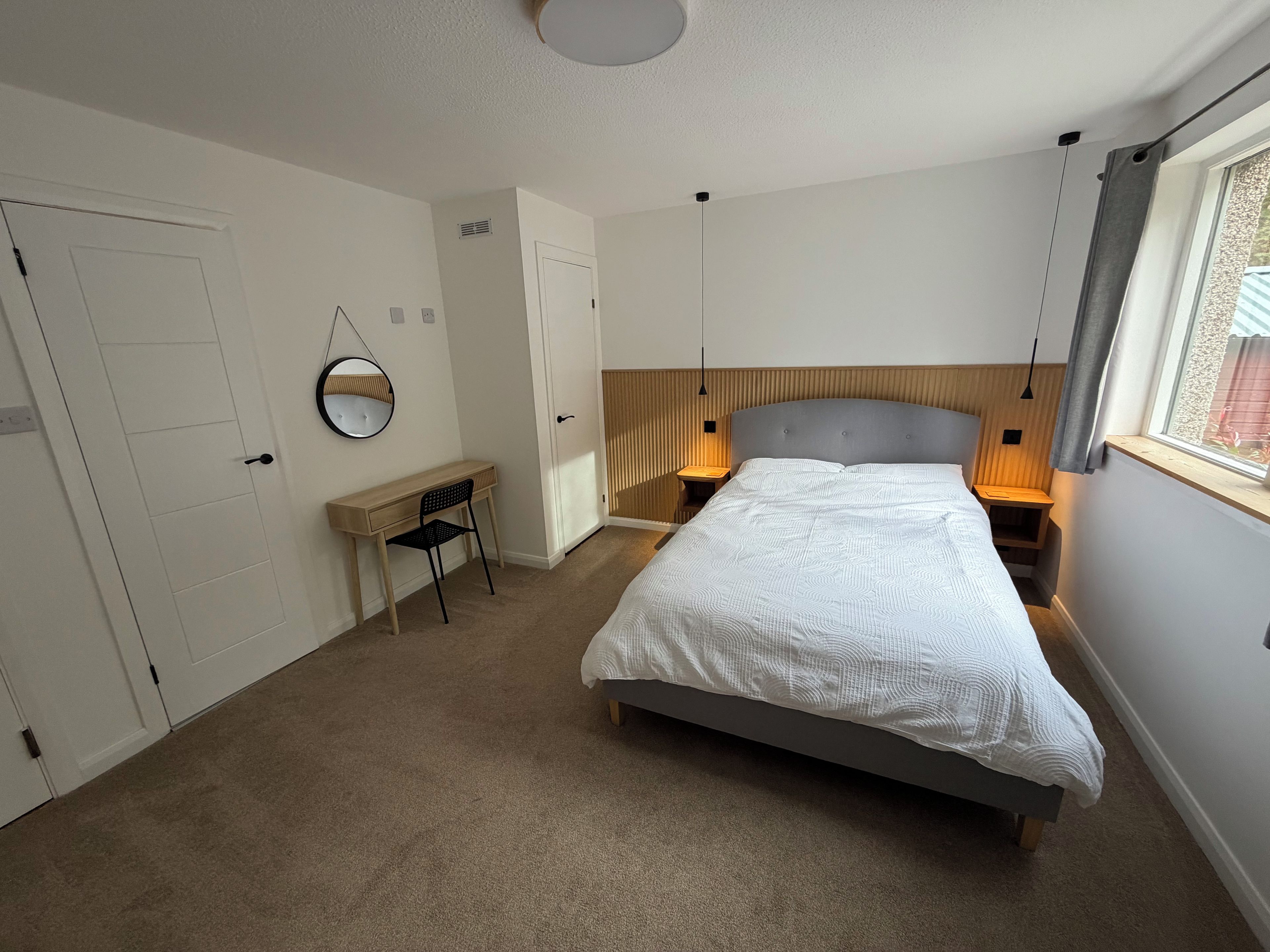 Ein Schlafzimmer mit Bett, Schreibtisch, Stuhl, Nachttisch, Lampe, Spiegel und Fenster.