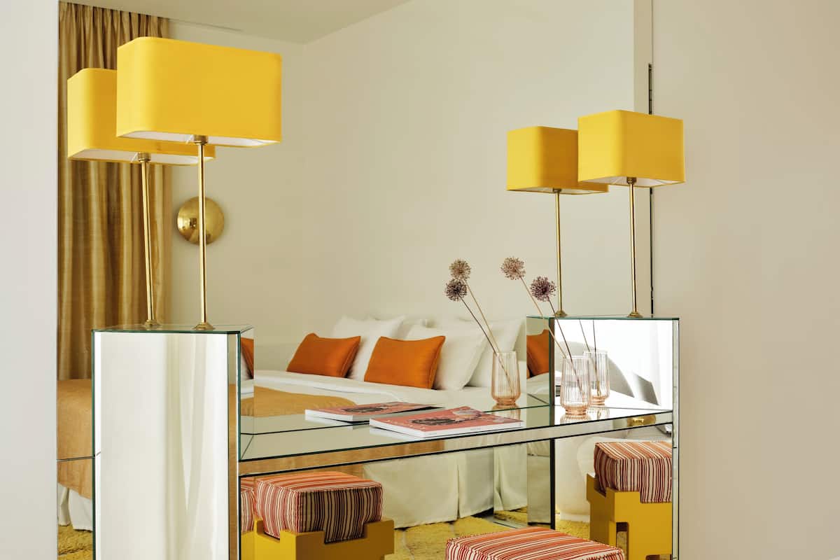 A modern hotel room with a glass coffee table, a sofa with orange cushions, and two yellow lamps.