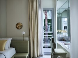 A modern hotel room with a green sofa, a small table with a lamp, and a view of a building through a glass door.