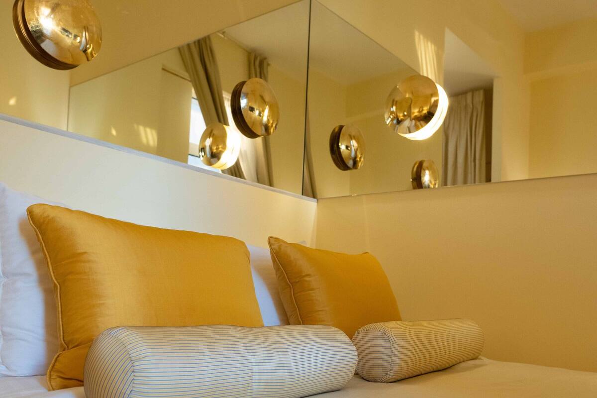 A neatly made bed with white and striped pillows, a large mirror above it, and a wall-mounted light fixture.