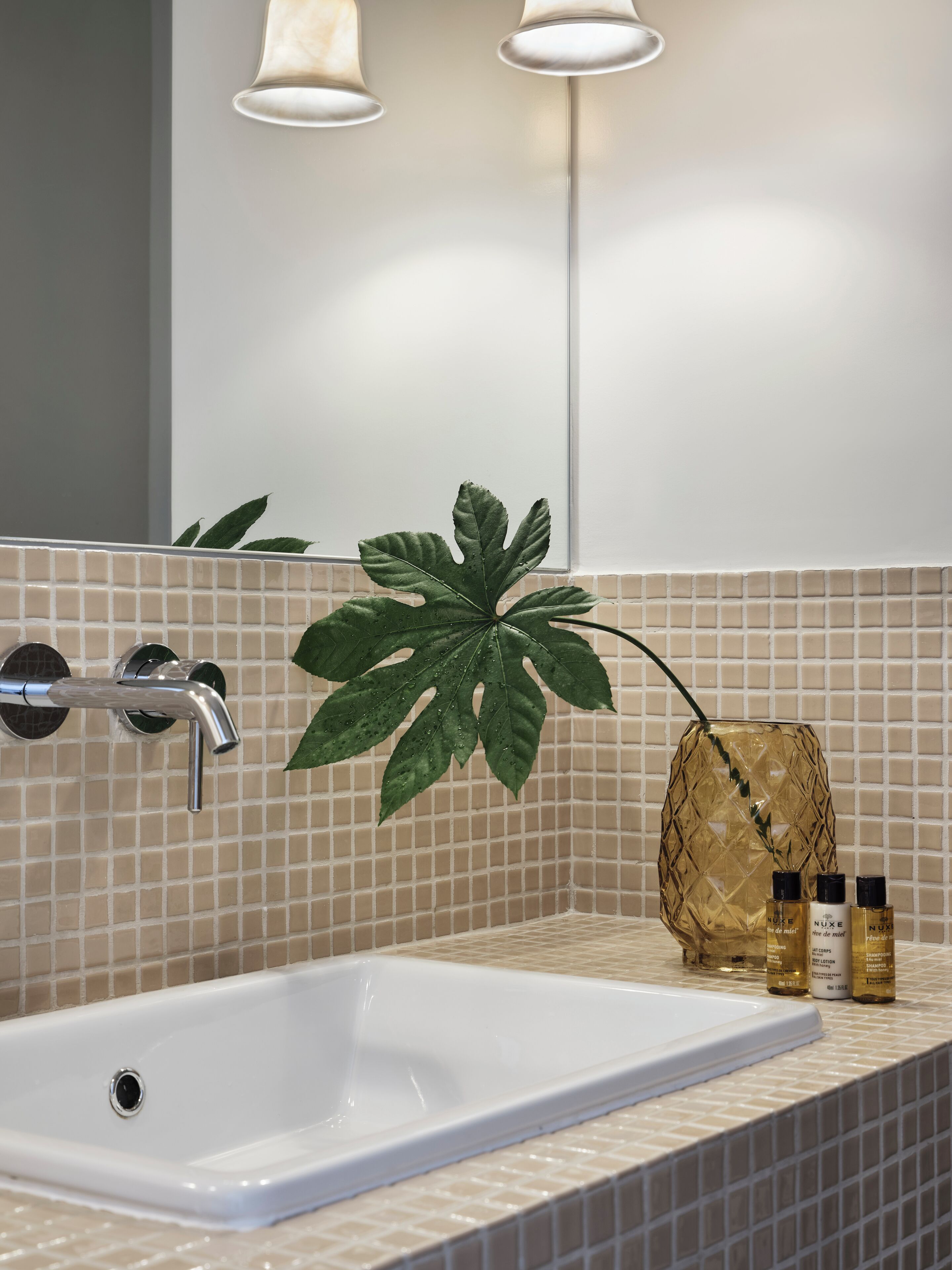 Ein Badezimmer mit einem weißen Waschbecken, einem Spiegel und einer Pflanze. Auf dem Waschtisch stehen zwei Shampoo-Flaschen und eine dekorative Vase.