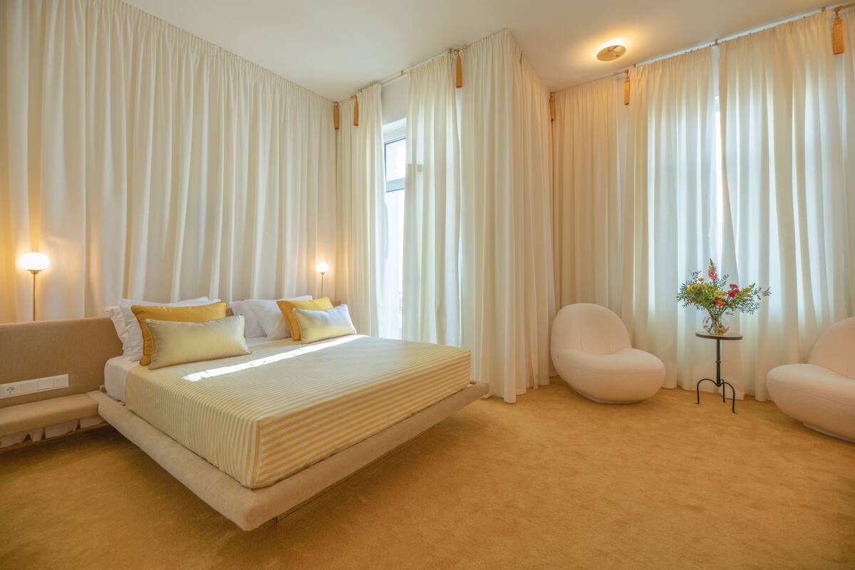 A spacious bedroom with a large bed, two chairs, a small table with a vase, and a bench.