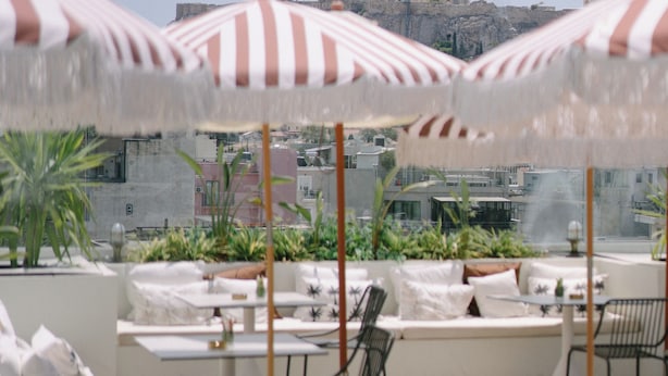 Een dakterras met gestreepte parasols, witte lounge fauteuils en een uitzicht op oude ruïnes.