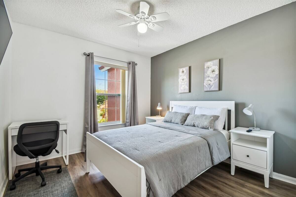 A bedroom with a bed, a desk, a chair, a nightstand with a lamp, and a ceiling fan.