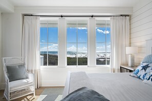 Une chambre avec une grande fenêtre, un lit, une chaise et une table de chevet avec une lampe.