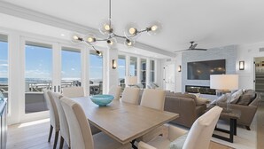 A spacious living room with a large TV, a dining area with a wooden table and chairs, and a view of the ocean through large windows. - PANORAMIC GULF FRONT in WATERSOUND BRIDGES! VIEWS, OUTDOOR BALCONY, 30A Area (Watersound)