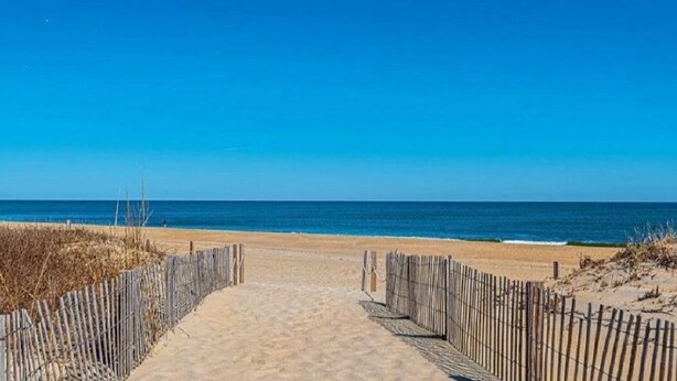 Een zandpad langs het strand dat naar de oceaan leidt, met een houten hek aan één kant.