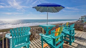 Une terrasse en bois avec des fauteuils Adirondack turquoise et un parasol bleu, donnant sur une plage.