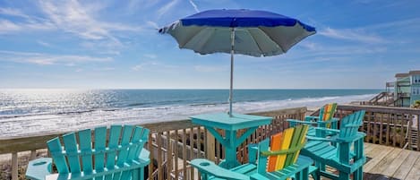 A wooden deck with turquoise Adirondack chairs and a blue umbrella overlooking a beach.